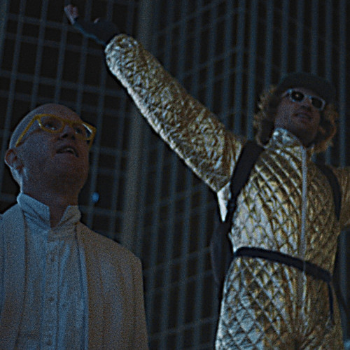 Man in gold suit with arms raised next to other man.
